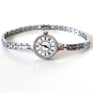 May include: A silver wristwatch with a white face, black Roman numerals, and blue floral details. The watch has a slim, linked bracelet-style strap. The watch is displayed on a white surface.