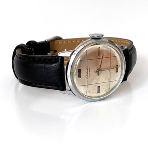 May include: Vintage Raketa watch with a silver-toned case and a black leather band. The watch face is a light beige color with black grid lines and the text "Raketa 21 Jewels". The watch has a classic, retro design.