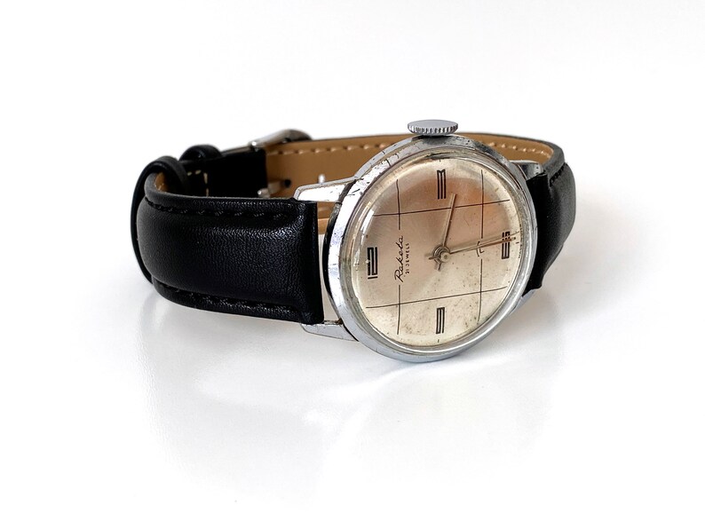 May include: Vintage Raketa watch with a silver-toned case and a black leather band. The watch face is a light beige color with black grid lines and the text "Raketa 21 Jewels". The watch has a classic, retro design.