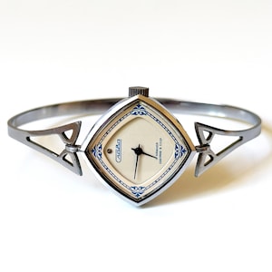 May include: A silver-toned bangle watch with a diamond-shaped case and a cream-colored dial. The watch face features blue decorative accents, black hands, and the text Slava in Cyrillic script. The band is a rigid, open-style metal cuff.