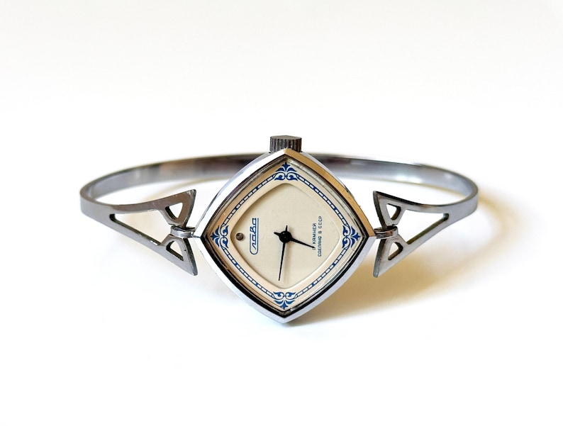 May include: A silver-toned bangle watch with a diamond-shaped case and a cream-colored dial. The watch face features blue decorative accents, black hands, and the text Slava in Cyrillic script. The band is a rigid, open-style metal cuff.