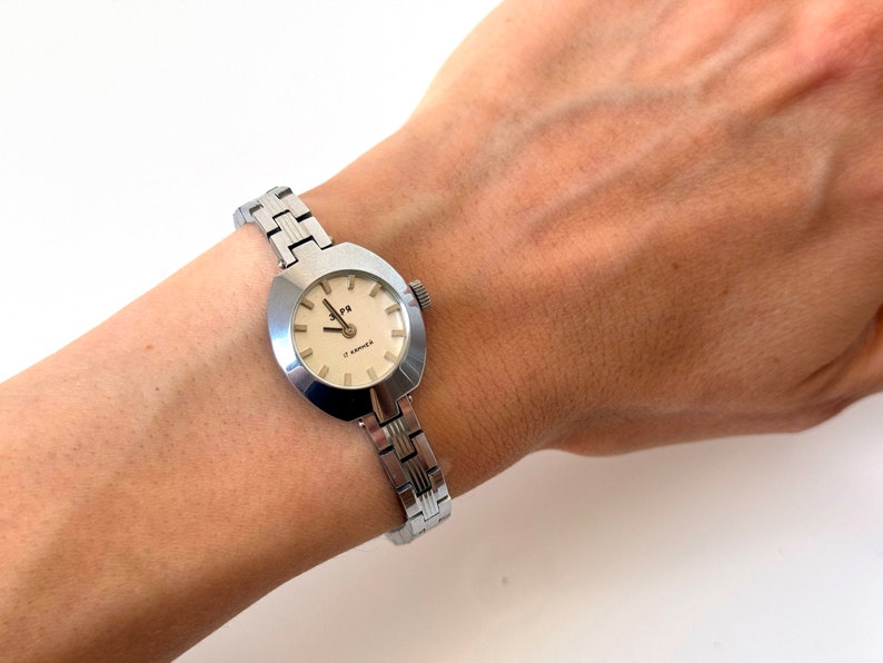 May include: A silver wristwatch with a round, slightly oval-shaped face. The watch face has a white background with black markings and the text "17 Jewels" and "C. KAMERER". The watch has a silver metal band.