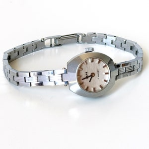May include: A silver wristwatch with a rectangular face and a silver metal strap. The watch face has a white background with black hour and minute hands and black markings for the numbers. The watch face also has the text "17 Jewels" printed on it.