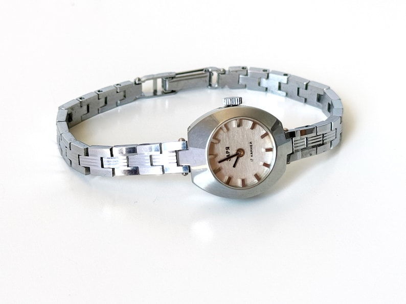 May include: A silver wristwatch with a rectangular face and a silver metal strap. The watch face has a white background with black hour and minute hands and black markings for the numbers. The watch face also has the text "17 Jewels" printed on it.
