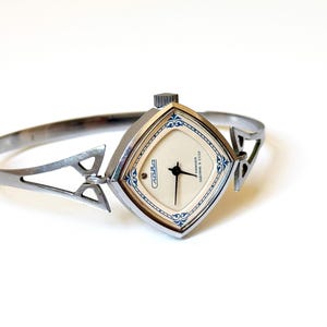 May include: A silver-toned bangle watch with a diamond-shaped case and a cream-colored dial. The watch face features black hands, blue decorative filigree accents at the corners, and the text Slava and Cdelano v CCCP printed in black.