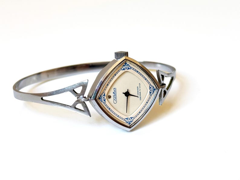 May include: A silver-toned bangle watch with a diamond-shaped case and a cream-colored dial. The watch face features black hands, blue decorative filigree accents at the corners, and the text Slava and Cdelano v CCCP printed in black.
