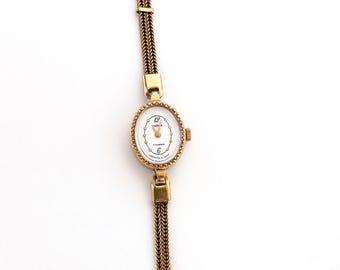 May include: A gold-tone wristwatch with an oval face, a white dial featuring black numerals at the 12 and 6 positions, and a textured gold-tone metal link bracelet. The watch case has a decorative scalloped edge and a small winding crown on the side.