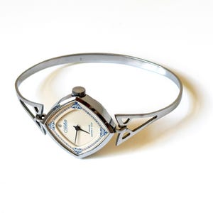 May include: A silver-toned bangle watch with a square-shaped case featuring rounded corners. The watch face is cream-colored with blue decorative accents, black hands, and the text Slava 17 jewels USSR printed on the dial. The band is a thin metal cuff.