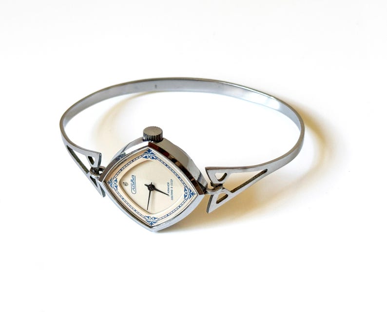 May include: A silver-toned bangle watch with a square-shaped case featuring rounded corners. The watch face is cream-colored with blue decorative accents, black hands, and the text Slava 17 jewels USSR printed on the dial. The band is a thin metal cuff.