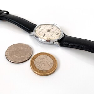 May include: A vintage wristwatch with a silver case and a black leather band. The watch face has a grid pattern and the word "Relide" is visible. A silver coin and a one Euro coin are next to the watch.