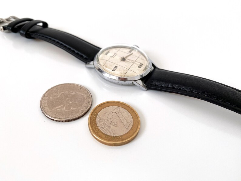 May include: A vintage wristwatch with a silver case and a black leather band. The watch face has a grid pattern and the word "Relide" is visible. A silver coin and a one Euro coin are next to the watch.