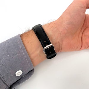 May include: A black leather watch strap with a silver buckle is fastened on a wrist. The watch strap is made of smooth, shiny black leather. The wrist is wearing a gray button-down shirt.