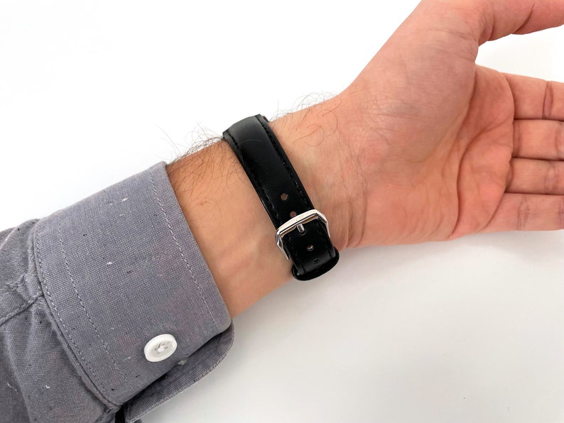May include: A black leather watch strap with a silver buckle is fastened on a wrist. The watch strap is made of smooth, shiny black leather. The wrist is wearing a gray button-down shirt.