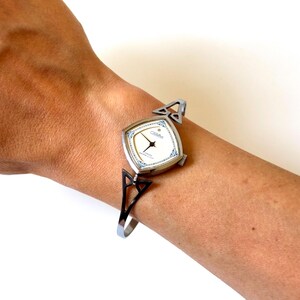 May include: A silver-tone wristwatch with a square face, white dial, and blue decorative accents. The watch features a thin, open-work metal bangle-style band with triangular cutouts on each side, connecting to the watch case with a simple crown.