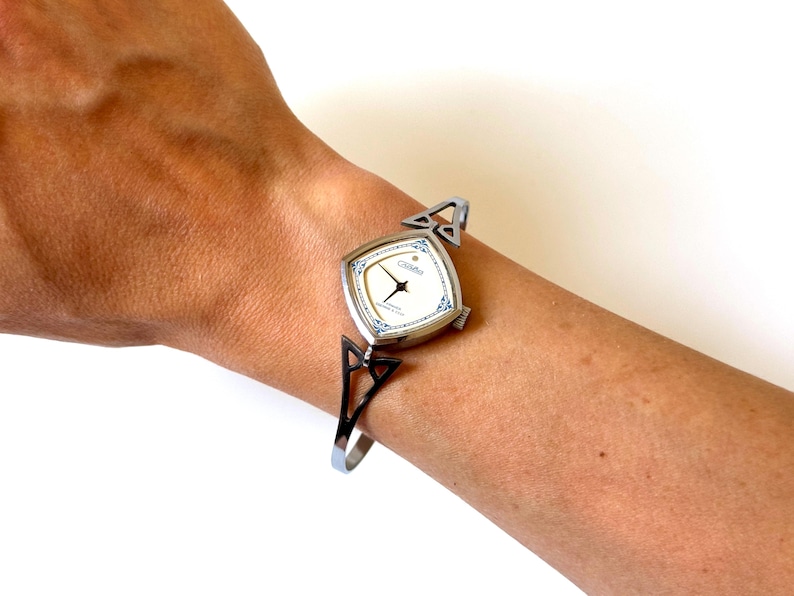 May include: A silver-tone wristwatch with a square face, white dial, and blue decorative accents. The watch features a thin, open-work metal bangle-style band with triangular cutouts on each side, connecting to the watch case with a simple crown.