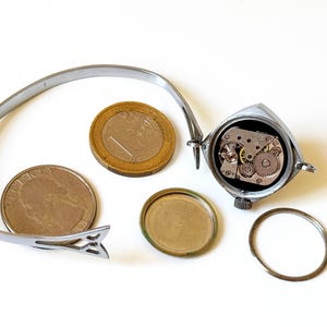 May include: A silver-toned wristwatch with an open back casing revealing the internal mechanical movement, accompanied by a detached circular back cover, a thin metal ring, and a flexible metal band, shown next to a one-euro coin and a US quarter.
