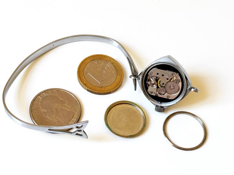 May include: A silver-toned wristwatch with an open back casing revealing the internal mechanical movement, accompanied by a detached circular back cover, a thin metal ring, and a flexible metal band, shown next to a one-euro coin and a US quarter.
