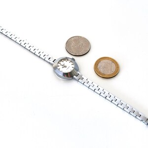May include: A silver wristwatch with a white face and a silver metal strap. The watch is next to two coins, one silver and one gold.