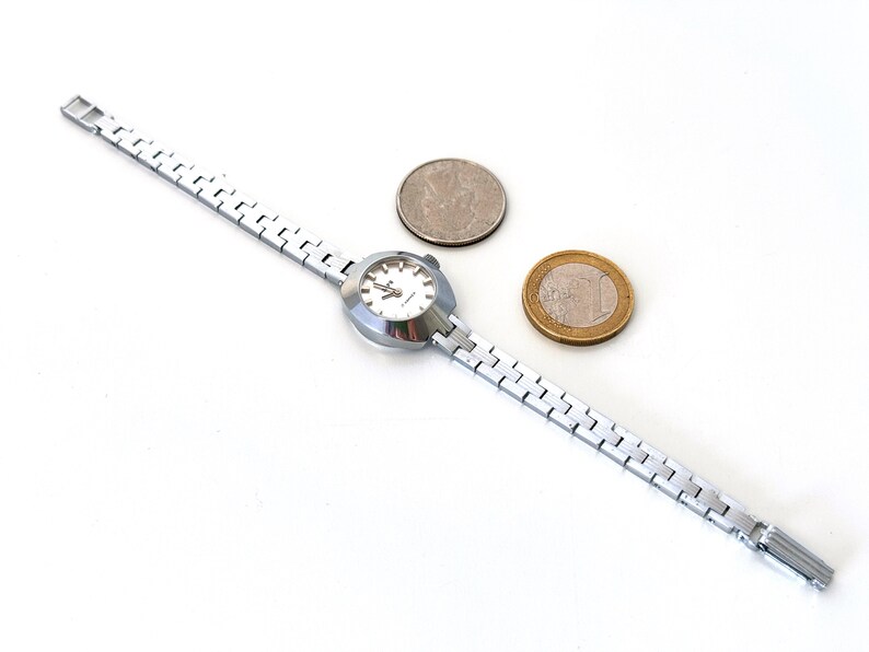 May include: A silver wristwatch with a white face and a silver metal strap. The watch is next to two coins, one silver and one gold.