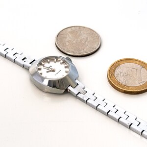 May include: A silver wristwatch with a white face and a silver metal band. The watch is next to a silver US quarter and a gold Euro coin.