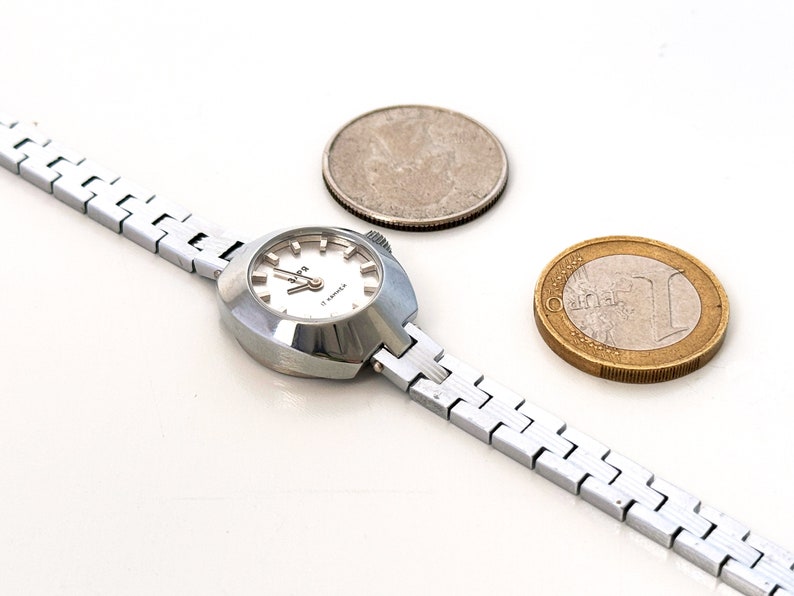 May include: A silver wristwatch with a white face and a silver metal band. The watch is next to a silver US quarter and a gold Euro coin.
