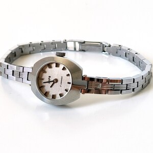 May include: A silver wristwatch with a silver metal strap. The watch face is oval shaped and has a white background with black markings. The watch face also has the text "3ПЧ" and "17 jewels" printed on it.