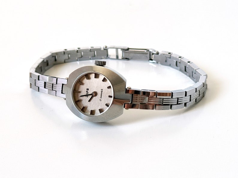 May include: A silver wristwatch with a silver metal strap. The watch face is oval shaped and has a white background with black markings. The watch face also has the text "3ПЧ" and "17 jewels" printed on it.