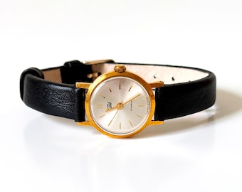 May include: A gold-tone wristwatch with a round silver dial, thin gold-tone hour markers, and gold-tone hands. The watch features a black leather strap with a gold-tone buckle. The dial includes a small logo and Cyrillic text near the bottom.