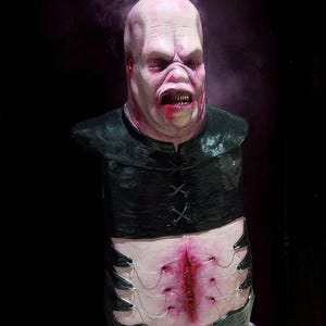 May include: A grotesque, pale-skinned bust with a bald head, snarling expression, and exposed teeth. The figure wears a black, leather-like top with criss-cross stitching. The lower torso features a gruesome, stitched wound, creating a horror-themed sculpture.