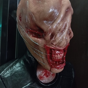 May include: A close-up of a horror-themed mask with a pale, fleshy complexion and a wide, gaping mouth. The mask has a series of wires running across the face, and the mouth is open, revealing sharp teeth and a bloody interior.