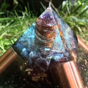 May include: A clear resin pyramid with copper accents and a copper coil at the centre. The pyramid contains blue and purple crystals and gold flakes. The pyramid is set against a backdrop of green grass.
