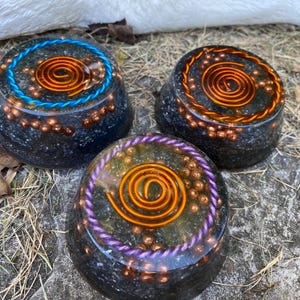 May include: Three round, black, resin-based, crystal healing orgonite bases with copper wire spirals and embedded crystals. Each base has a different coloured wire wrap around the edge: blue, purple, and orange.
