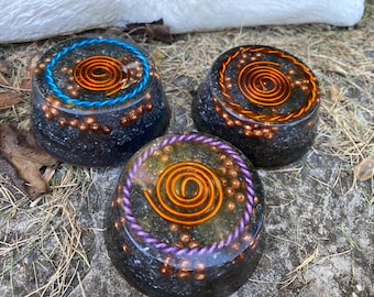 Può includere: Tre basi rotonde in orgonite di guarigione dei cristalli, a base di resina nera, con spirali di filo di rame e cristalli incorporati. Ogni base ha un filo di colore diverso attorno al bordo: blu, viola e arancione.