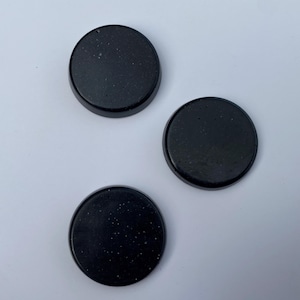 May include: Three black, circular coasters with a speckled texture. The coasters have a raised edge and a smooth surface. They are arranged on a white background.