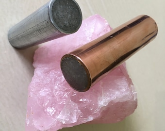 Puede incluir: Dos varillas metálicas, una plateada y otra de cobre, se colocan sobre un gran cristal de cuarzo rosa. La varilla plateada tiene una superficie texturizada y la varilla de cobre tiene una superficie lisa.