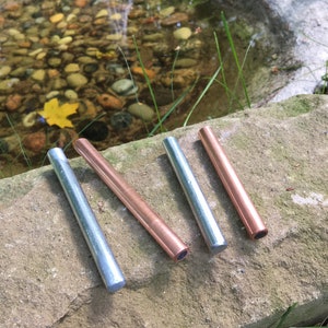 May include: Four metal tubes, two silver and two copper, are arranged diagonally on a stone surface. The tubes are cylindrical with a flat end. The background includes a small pond with rocks and vegetation.