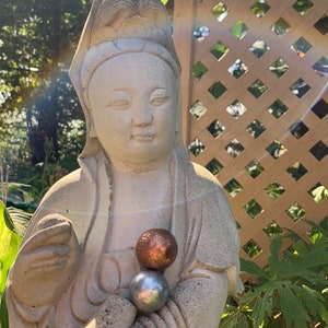Puede incluir: Estatua de piedra desgastada de una figura serena, posiblemente una deidad, con una pequeña escultura en la cabeza. La estatua sostiene dos esferas, una de cobre y otra de plata. El fondo incluye un enrejado de madera y exuberante vegetación.