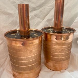 Peut inclure: Deux tasses en cuivre avec un tuyau en cuivre qui s'étend du haut. Les tasses ont une finition martelée et un bord de couleur or.