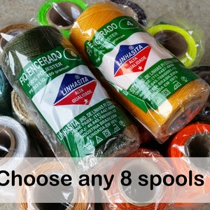 Pode incluir: Um close-up de uma variedade de carretéis de linha em diferentes cores, incluindo verde, amarelo, laranja, branco e preto. Os carretéis são etiquetados com "FIO ENCERADO" e "LINHASITA ALTA QUALIDADE". Escolha 8 carretéis!
