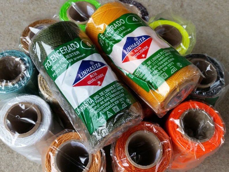 May include: A collection of spools of waxed polyester thread in various colors, including green, orange, yellow, and white. The spools are labeled with the brand name "LINHASITA" and the text "FIO ENCERADO". Each spool contains 100 grams of thread.