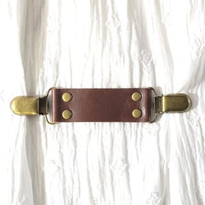 May include: A brown leather strap with two metal clips and four brass rivets. The strap is on a white fabric background.