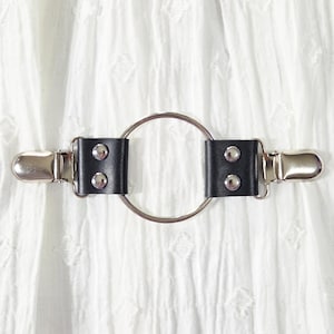 May include: A black leather garter belt with a silver metal ring and two silver metal clips. The garter belt has two black leather rectangles with two silver rivets each. The background is a white, textured fabric.