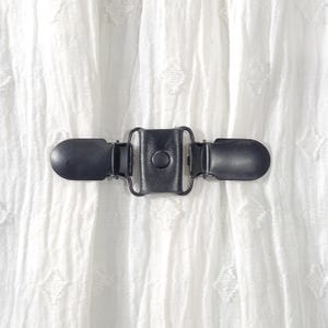 May include: A black metal hook and eye closure with a round button in the center. The closure is on a white fabric background.