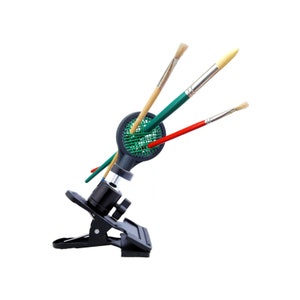 Puede incluir: Un soporte negro y verde para pinceles con tres pinceles. El soporte tiene una base de clip y un cuello flexible. Los pinceles tienen mangos de madera y cerdas de varios colores. El fondo es blanco.