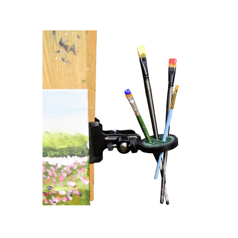 Puede incluir: Un soporte negro y ajustable para pinceles de arte, fijado a un caballete de madera. El soporte contiene varios pinceles con cerdas coloridas. Tambi&eacute;n se ve un peque&ntilde;o lienzo con una pintura de paisaje.