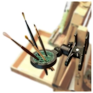 Puede incluir: Un soporte negro para pinceles de artista con una pinza, que sostiene varios pinceles. El soporte tiene un dise&ntilde;o circular con una inserci&oacute;n de malla verde para sujetar los pinceles. La pinza permite fijar el soporte a una mesa o caballete.