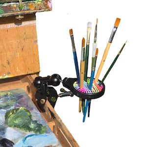 May include: A black, adjustable paint brush holder with a clamp, attached to a wooden easel. The holder contains various paint brushes with wooden handles and colorful bristles. The easel has a palette with visible paint.