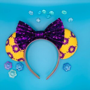 May include: A headband with yellow ears featuring a purple floral pattern. The headband has a large purple sequined bow. The headband is on a blue background with decorative blue and purple flower-shaped embellishments.