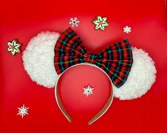 May include: A festive headband with white fluffy ears, a plaid bow, and a red interior. The bow features a pattern of red, green, blue, and yellow. The headband is decorated with snowflake and gingerbread man accents, all set against a vibrant red background.