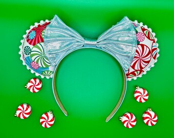 May include: A pair of light blue Minnie Mouse ears with a silver bow and a peppermint candy cane pattern. The ears are decorated with white pom-poms. There are also several small peppermint candy cane ornaments scattered around the ears.
